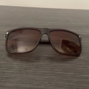Bruna Rayban liknande solglasögon - Snygga solglasögon med bruna bågar och fyrkantig form. Glasen är tonade i brunt och bågarna har en stilren, enkel design. Perfekta för att ge en cool vibe till din outfit. Liten tecken på användning därframme men inte mycket som syns till. Priset är hugget i stenen! Men kan sänkas i snabb affär 