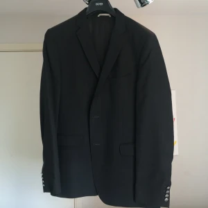 Svart kavaj från BOSS med silvriga knappar - Stilren svart kavaj från BOSS med klassisk passform och snygga silvriga knappar framtill och vid ärmslut. Kavajen har två fickor med lock, bröstficka och helfodrad insida. Perfekt för dig som vill ha en tidlös och elegant look.