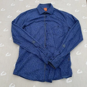 Blå mönstrad skjorta från Hugo Boss - Snygg blå skjorta från Hugo Boss i slim fit med smått mönster i vitt och svart. Skjortan har klassisk krage, bröstficka och knäppning framtill. Perfekt för dig som gillar stilrena detaljer och en smalare passform.