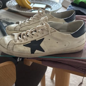 Golden Goose Superstar sneakers vit/svart - Golden Goose Superstar sneakers i vitt skinn med svarta detaljer och ikonisk stjärna på sidan. Klassisk låg modell med snörning och platt sula. Bakpartiet är svart med vit logga. Perfekta för en cool och avslappnad streetstil.