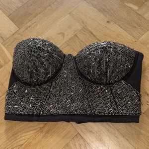 Svart glittrig korsettopp från Bik Bok - Supersnygg svart korsettopp från Bik Bok med glittriga silverdetaljer och geometriskt mönster framtill. Toppen är axelbandslös och har dragkedja bak för enkel påklädning. Perfekt för att sticka ut och ge din outfit extra edge.