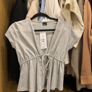 Grå tröja med knyt från Gina Tricot - Supersöt ljusgrå omlottopp från Gina Tricot med korta ärmar och knyt i midjan. Toppen har en v-ringning och är tillverkad i mjuk bomullsmix. Passar S och XS