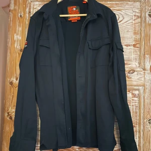 Svart overshirt från Superdry - Säljer en svart overshirt från Superdry Rookie Edition i storlek 2XL, MEN sitter som en L. Skjortan har två bröstfickor med lock, dragkedja och knappar framtill samt coola detaljer på ärmen. Knappt använd