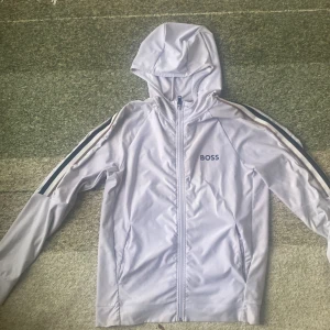 HUGO BOSS tröja - ÄKTA Snygg vit vindjacka från BOSS med huva och hel dragkedja. Jackan har svarta och vita ränder längs ärmarna och BOSS-logga på bröstet. Tillverkad i polyester, perfekt för dig som gillar sportig och clean stil.