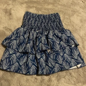 Blå volangkjol med mönster - Supersöt blå kortkjol med vitt grafiskt mönster och bred resår i midjan. Kjolen har flera volanger som ger en riktigt fin look. Perfekt för varma sommar kvällar