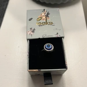 Blå evil eye-ring från Ioaku - Cool ring från Ioaku med ett blått evil eye-motiv i mitten, omgiven av små glittrande stenar. Ringen har en rund form och silverfärgad bas. Perfekt statement piece för dig som gillar unika accessoarer.