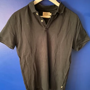 Svart piké från East West i bomull - Svart pikétröja från East West i ekologisk bomull. Klassisk krage, kortärmad och med knappslå framtill. Enkel och stilren design som funkar till det mesta. Perfekt för dig som gillar en clean och tidlös look.