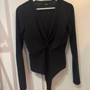 Svart omlott bodysuit från Bik Bok - Svart långärmad bodysuit från Bik Bok med djup v-ringning och omlott-twist framtill. Tight passform och mjukt stretchigt material som sitter snyggt på kroppen. Perfekt till jeans eller kjol för en trendig look.