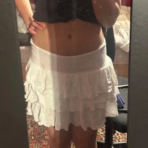 Vit volangkjol med resårmidja - Supersöt vit kortkjol med flera lager volanger och bred smockad resår i midjan. Kjolen är luftig och lätt, perfekt för varma dagar. Märke STR, passar dig som gillar en feminin och trendig stil.