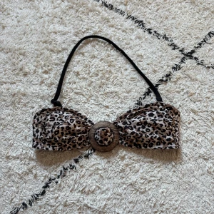Leopardmönstrad bikiniöverdel Lindex - Snygg bikiniöverdel från Lindex i leopardmönster med bruna och svarta toner. Bandeau-modell med avtagbar axelrem och coolt träinspirerat spänne framtill. Perfekt för strandhäng och poolparty.
