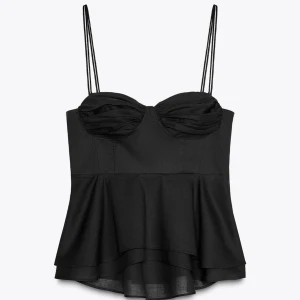 Svart volangig korsettopp - Supersnygg svart korsettopp med dubbla tunna axelband och volangig nederdel. Toppen har draperade kupor och en figurnära passform.Perfekt för dig som vill sticka ut med en cool och feminin look.
