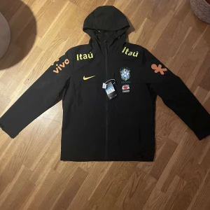 Nike Brasil svart vindjacka - Svart Nike vindjacka med huva och dragkedja, Brasil-logga och sponsormärken i gult och orange. Jackan har lång ärm, snygga tryck och är tillverkad i lätt och ventilerande syntetmaterial med Dri-FIT-teknologi. Perfekt för dig som älskar fotboll och vill ha en sportig look.