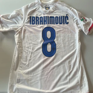 Inter Milan Ibrahimović tröja Nike - Inter Milan bortatröja med Ibrahimović och nummer 8 på ryggen. Vit bas med rött kors på framsidan, blå detaljer och klubbmärke. Nike-logga och Pirelli-sponsor framtill. Tillverkad i lätt och ventilerande material, perfekt för fotbollsfans. Replika. Endast handtvätt gäller!