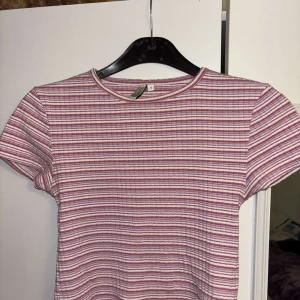 Randig ribbad topp från NLY Trend - Superfin topp från NLY Trend i storlek S. Den är ribbad och har ett randigt mönster i rosa och vitt. Modellen är kortärmad med rund halsringning.