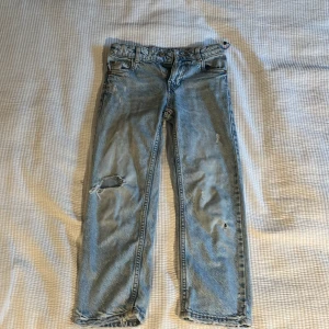 Ljusblå slitna jeans från Zara - Ljusblå jeans från Zara med raka ben, slitna detaljer och hål på ena benet. Jeansen har fransade benslut och klassisk femficksdesign. Perfekta för en avslappnad och trendig look.