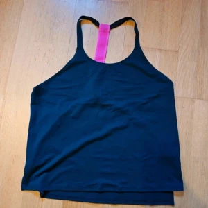 Svart träningstopp Fabletics Demi Lovato - Sportig svart linnetopp från Fabletics Demi Lovato-kollektionen. Toppen har en cool rosa meshdetalj i ryggen och smala axelband. Perfekt för träning och gjord i stretchigt material som andas, 88% polyester och 12% elastan.