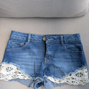 Jeansshorts med spets från Clockhouse - Blå lågmidjade jeansshorts från Clockhouse med snygga slitna kanter och vit spetsdetalj längs bensluten. Stl 34