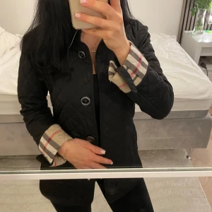 Svart quiltad jacka från Burberry S (äkta)  - Säljer en svart quiltad jacka från Burberry med klassiskt rutigt foder i beige, rött och svart. Jackan har knappar framtill, långa ärmar och snygga uppvikta muddar som visar det ikoniska mönstret. 