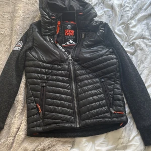 Svart Superdry Storm pufferjacka M - Svart pufferjacka från Superdry Storm med quiltad front och huva. Jackan har gråmelerade ärmar, två dragkedjefickor och coola orange detaljer. Perfekt för dig som vill ha en sportig och urban look med snygga kontraster och tydlig logga på ärmen.