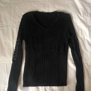 Svart ribbad stickad tröja med text - Svart ribbad stickad tröja med v-ringning och långa ärmar. Ena ärmen har texten 'ANGEL' i silvriga nitar och den andra 'DEVIL'. Tröjan har en tight passform men är väldigt stretchig. Märket heter prodotto italiano.