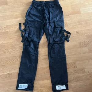 Svarta cargopants från Sixth June S - Svarta cargopants från Sixth June med elastisk midja och snörning. Byxorna har flera stora fickor, coola banddetaljer på sidorna och patchar vid bensluten. Perfekta för en streetwear-look och riktigt bekväma med loose fit.