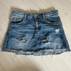 Blå jeanskjol från Zara med slitningar - Säljer en blå jeanskjol från Zara med rå kant och slitna detaljer framtill. Kjolen har klassisk femficksdesign, knapp och dragkedja i midjan samt broderad logga bak. Perfekt för dig som gillar en avslappnad och trendig stil.