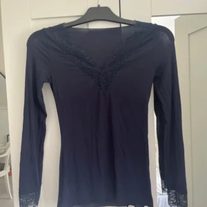 Mörkblå långärmad topp med spets - Supersnygg mörkblå långärmad topp med v-ringning och spetsdetaljer både vid halsen och ärmsluten. Tunn och mjuk i materialet, perfekt för lager-på-lager eller som den är. Sitter tight och har en feminin vibe med spetsen som extra detalj.