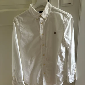 Vit skjorta från Ralph Lauren - Klassisk vit skjorta från Ralph Lauren med broderad logga på bröstet. Skjortan har lång ärm, knappar hela vägen och en snygg krage. Modellen är custom fit och tillverkad i bomull, perfekt för en clean och stilren look. 