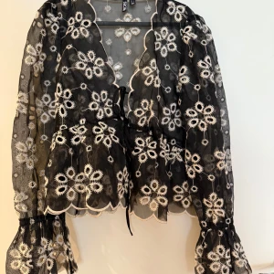 Svart blommig blus med broderi - Superfin svart transparent blus  med vita broderade blommor, volangdetaljer och knyt framtill. Långa puffiga ärmar med resår vid handlederna och vågig kant nertill. Perfekt för dig som gillar unika detaljer och romantisk stil.