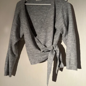 Grå omlottkofta med knytband S - Mysig grå omlottkofta med långa ärmar och breda knytband i midjan. Koftan är stickad och har en djup v-ringning, perfekt att slänga på sig över en topp. Enkel och stilren design som passar till många outfits.