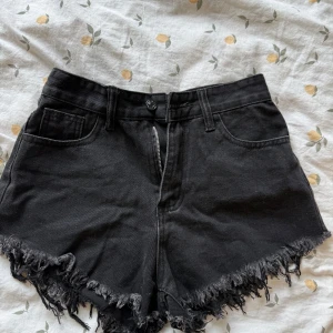 Svarta jeansshorts med flames - Svarta högmidjade jeansshorts med fransig kant och coola vita flames broderade på bakfickorna. Klassisk femficksmodell med dragkedja och knapp. Perfekta för en edgy sommarlook.