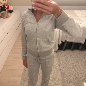 Grå Juicy Couture med strass - Supermysiga grått sett från Juicy Couture med strassdetaljer längs benet och bakfickor med knappar. Byxorna har dragsko i midjan och är tillverkade i mjukt material som känns skönt mot huden. Perfekta för chill dagar hemma eller när du vill vara extra comfy.