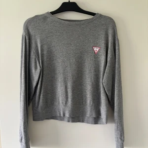 Grå sweatshirt från Guess i bomull - Säljer en stilren grå sweatshirt från Guess i mjuk bomull. Tröjan har lång ärm, rund halsringning och en liten rödvit Guess-logga på bröstet. Perfekt basic att ha i garderoben och lätt att matcha med jeans eller kjol.