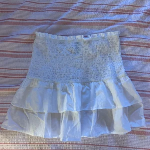 Vit volangkjol från H&M Divided - Supersöt vit kortkjol från H&M Divided i storlek XXS. Kjolen har smockad midja och dubbla volanger som ger en luftig och somrig känsla. Perfekt för dig som gillar en feminin och trendig stil.
