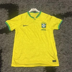 Brasilien Nike fotbollströja gul - Gul Brasilien fotbollströja från Nike med gröna och blå detaljer på krage och ärmslut. Broderad CBF-logga och Nike swoosh på bröstet. Knappar vid halsen med brasiliansk flaggdetalj. Tillverkad i lätt och ventilerande polyester. Priset är såklart inte hugget i sten!