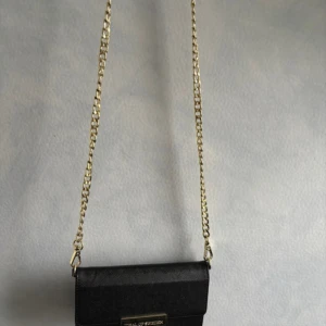 Svart mobil och axelväska från iDeal of Sweden - Snygg svart mobilskal med  axelväska från iDeal of Sweden med guldfärgad kedja och metallogo framtill. Väskan har stilren design, flera kortfack på insidan och är gjord i strukturerat syntetmaterial. Perfekt för att bära det viktigaste med stil.