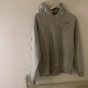 Grå hoodie med stjärnor från Gina Tricot - Grå hoodie från Gina Tricot med svart text 'MA CHERIE' och en stjärna framtill. På båda ärmarna finns flera svarta stjärnor tryckta längs hela ärmen. Tröjan har huva och är långärmad, säljer pga att den blivit för liten. 