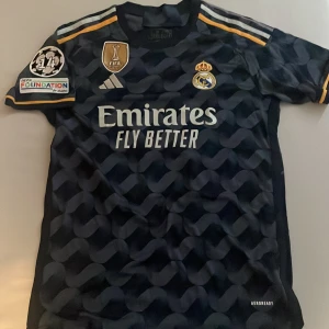 Real Madrid bortatröja Tchouameni 18 - Mörkblå Real Madrid bortatröja med Tchouameni 18 på ryggen. Tröjan har korta ärmar, guldiga detaljer och mönster över hela tyget. Broderad klubbmärke, Adidas-logga och sponsortryck framtill. UEFA Foundation och FIFA World Champions 2022 patchar på ärmen. Äkta och nyskick. Ingår även real madrid #camavinga.