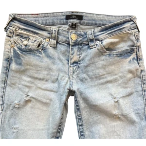 Ljusblå jeansbyxor från True Religion med slitningar  - Säljer ett par ljusblå jeans från True Religion med slitna detaljer och klassisk broderad bakficka. Byxorna har låg midja, med bootcut modell. Jeansen har två snygga hål vid knäna. Säljer då de är för korta för mig som är 168. Kan skicka fler bilder med dem på🙏🤍Storleken är w27