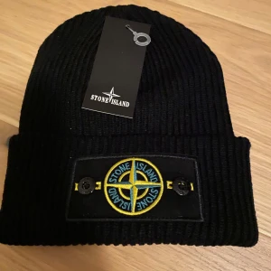 Svart ribbstickad mössa från Stone Island - Svart ribbstickad mössa från Stone Island med uppvikt kant och klassisk logotyp-patch framtill. Märket är broderat i gult, blått och grönt. Perfekt accessoar för kyliga dagar.
