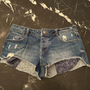 Jeansshorts med paisleydetalj - Snygga blå jeansshorts med slitningar och råa kanter. Insidan av benen har ett mörkblått paisleymönstrat tyg som sticker fram. 