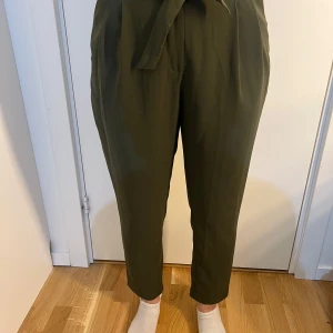 Olivgröna kostymbyxor med knytband - Snygga olivgröna kostymbyxor med hög midja och knytband i midjan. Byxorna har en rak passform och pressveck framtill, samt diskreta fickor bak. Perfekta för en stilren och trendig look.