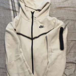 Vit hoodiejacka från Nike i storlek XS passar S - Snygg vit hoodiejacka från Nike med svart dragkedja och logga på bröstet. Jackan har huva, lång ärm och en extra ficka med dragkedja på ena ärmen. Perfekt för en sportig look.