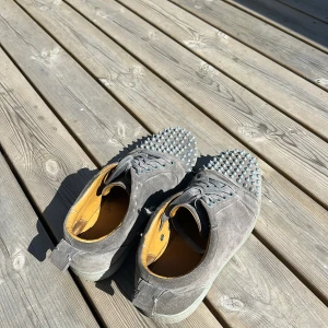 Grå mockasneakers med nitar från Christian Louboutin - Säljer ett par grå sneakers i mocka från Christian Louboutin med ikoniska röda sulor och silverfärgade nitar på tån. Skorna har snörning och en rund tå. Insidan är ljusgul och sulan är platt. Fick dem här på jul klapp 2023 utan kvittot. 