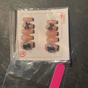 Falska naglar med svart dekor och tillbehör - Säljer ett set med lösnaglar i nude färg med svart kant och dekorationer som rosetter och fyrkanter. Paketet innehåller även självhäftande klisterlappar och en rosa nagelfil. Perfekt för att fixa snygga naglar snabbt hemma.