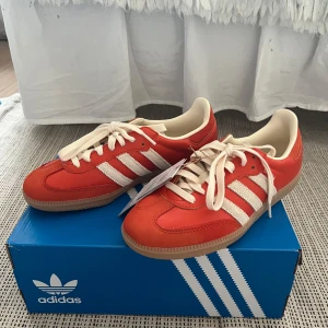 Helt NYA Adidas Samba OG i orange och vitt - Säljer ett par Adidas Samba sneakers i orange-röd med krämvita detaljer och beige sula. Klassisk modell med snörning och retrovibbar. Materialet är både mocka och läder vilket ger en cool och unik look. Skorna är helt nya och aldrig använda, har provat dem en gång inomhus. Nypris är 1349kr på ASOS. 