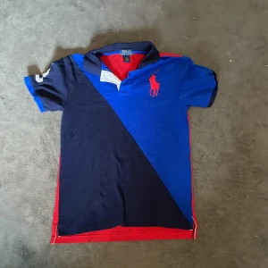 Blå och marinblå pikétröja från Polo Ralph Lauren - Säljer en snygg pikétröja från Polo Ralph Lauren i blått och marinblått med röd logga på bröstet. Tröjan har krage, korta ärmar och röda detaljer längs sidorna. Tillverkad i 100% bomull.