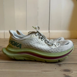 Vita löparskor från Hoka med grön sula modell Kawana - Säljer ett par vita Hoka löparskor med ventilerande mesh-ovandel och tjock, grön mellansula med rosa detalj. Skorna har snörning och en bred, stabil platt sula som ger bra dämpning. Perfekta för dig som gillar att springa långt och vill ha maximal komfort.