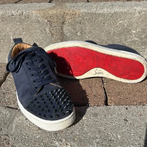 Mörkblå sneakers med nitar från Christian Louboutin - Säljer ett par mörkblå sneakers från Christian Louboutin med nitar på tån och den klassiska röda sulan. Skorna har snörning och är tillverkade i mocka med vita gummisulor. Perfekta för dig som vill sticka ut med din stil. Skorna saknar en av nitarna som sitter på (därav priset) men det är inget som syns då den spiken som saknas sitter på vänster sko på insidan längst in och längst ner. Kvitto finns EJ! Om det är något mer du undrar är det bara att skriva mvh Otto!😃