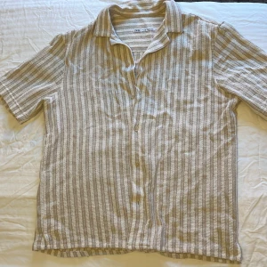 Beige och vit randig skjorta från Zara - Säljer en kortärmad skjorta från Zara i beige och vitt med diskret randigt mönster. Skjortan har klassisk krage och knäppning framtill. Perfekt för varma dagar och enkel att styla med jeans eller shorts.